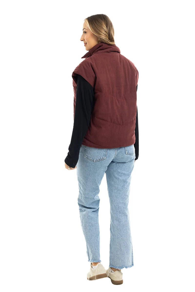 A woman models the Katydid Wine Fall Corduroy Puffer Vest over a black long-sleeve shirt, light blue jeans, and beige shoes. Her hair is down as she faces away against a white background—ideal trendy outerwear for autumn.