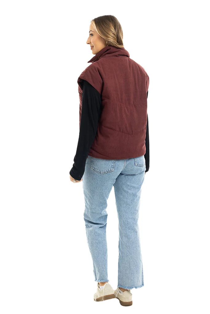 A woman models the Katydid Wine Fall Corduroy Puffer Vest over a black long-sleeve shirt, light blue jeans, and beige shoes. Her hair is down as she faces away against a white background—ideal trendy outerwear for autumn.