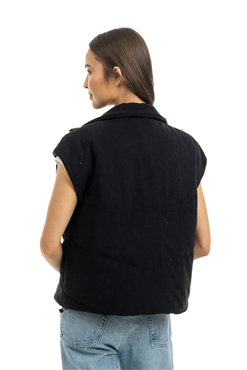 A person with long brown hair stands facing away from the camera in light blue jeans and a Katydid Black Corduroy Trendy Puffer Vest against a white background.