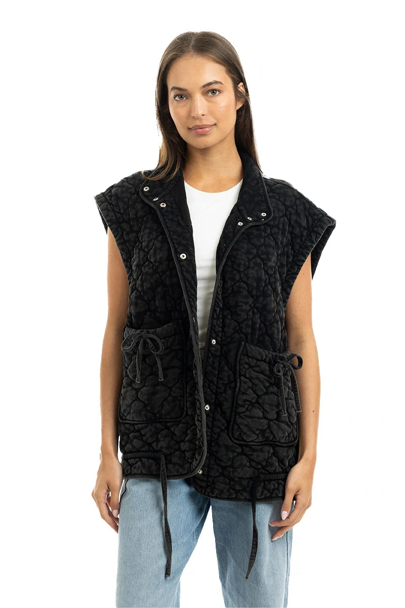 A woman wears the Katydid Charcoal Oversized Garment Washed Quilted Vest with front pockets and tie details over a white shirt and light blue jeans, standing against a white background.
