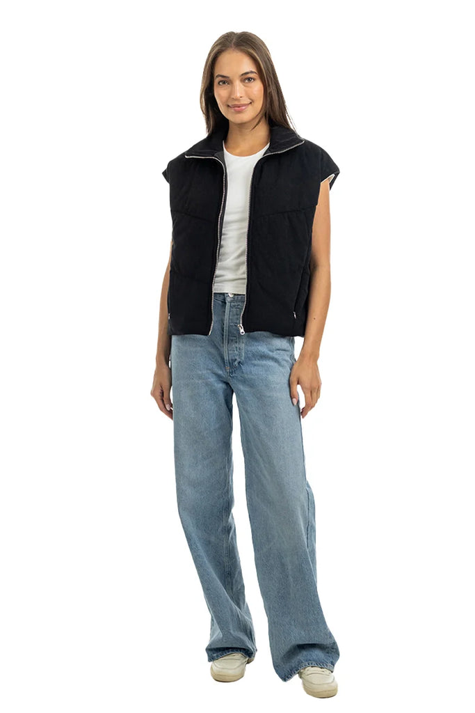 A woman with long brown hair smiles against a white background, wearing the Katydid Black Corduroy Trendy Puffer Vest over a white t-shirt, paired with light blue wide-leg jeans and white sneakers.