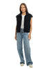 A woman with long brown hair smiles against a white background, wearing the Katydid Black Corduroy Trendy Puffer Vest over a white t-shirt, paired with light blue wide-leg jeans and white sneakers.