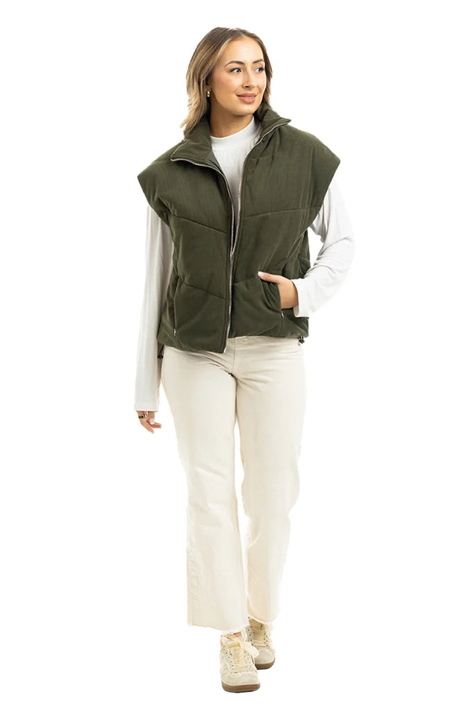 A person wears the Katydid Olive Corduroy Puffer Vest over a long-sleeve white shirt, paired with off-white pants and beige shoes. Standing against a white background, they smile slightly to the side with one hand in their pocket.