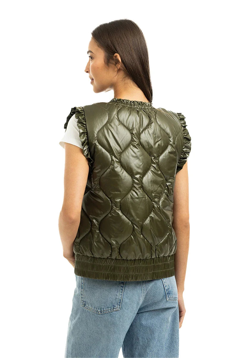 A woman with straight brown hair, facing away from the camera, wears the Katydid Olive Metallic Puffer Lightweight Vest over a white top and light blue jeans.