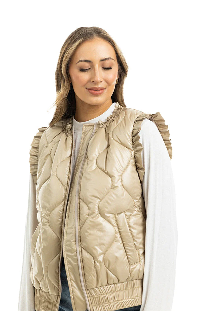 A woman smiles with her eyes closed against a white background, modeling the Katydid Soft Tan Metallic Quilted Vest with ruffled shoulders over a white long-sleeve shirt, highlighting the vest’s stylish charm.