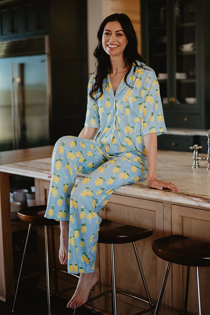A woman with long dark hair wears the Katydid Zesty Lemons Matching Pajama Pants Set, featuring a blue fabric with yellow lemon print, as she sits barefoot on a kitchen counter and smiles at the camera.