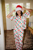 A woman in Katydid’s Santas Mail Christmas Pajamas for Women and a Santa hat smiles as she poses indoors on a brick floor near glass doors.