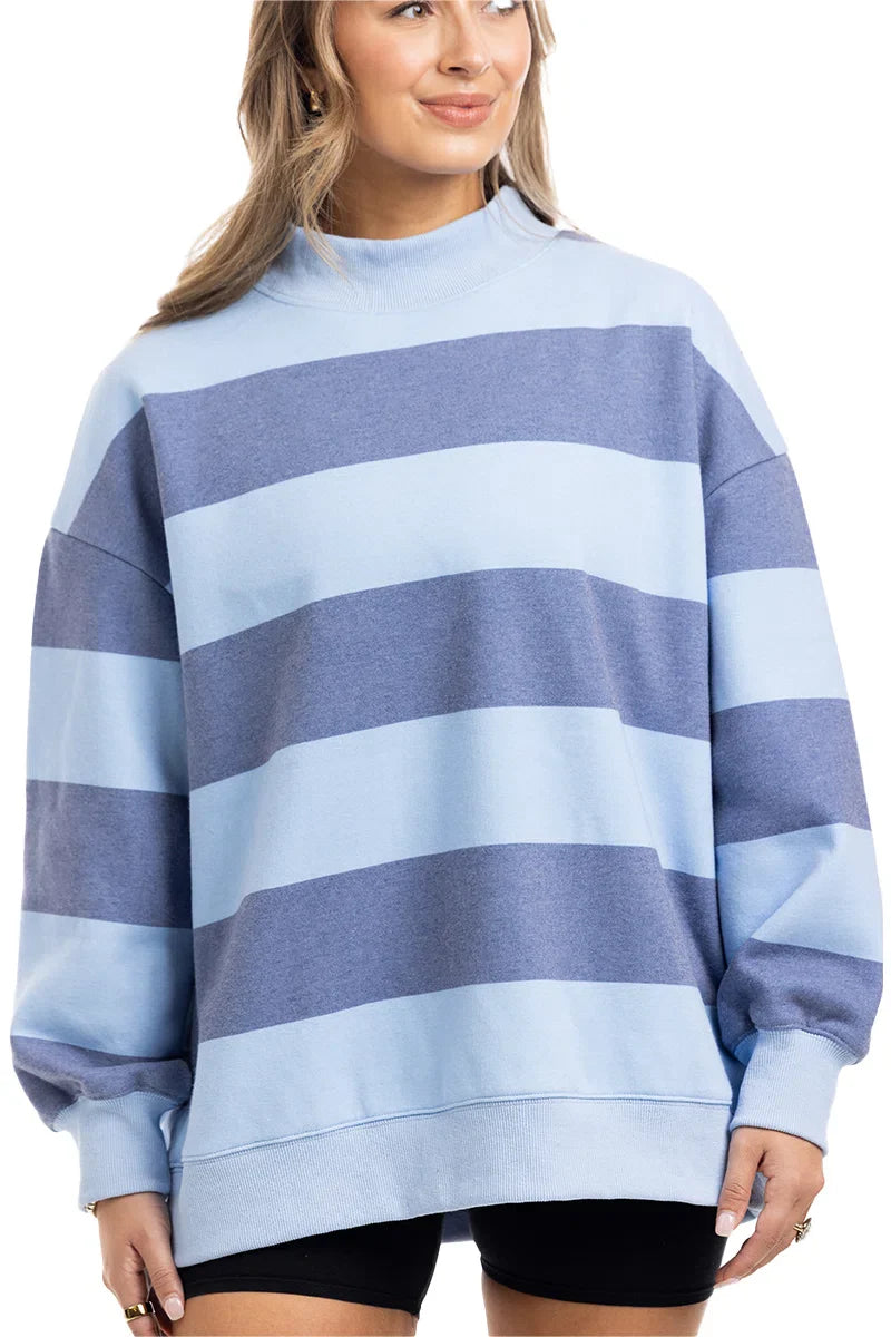 A person stands smiling with their hair down, wearing the Katydid Light Blue and Navy Striped Mockneck Sweatshirt—perfect for fall layering—and black shorts.