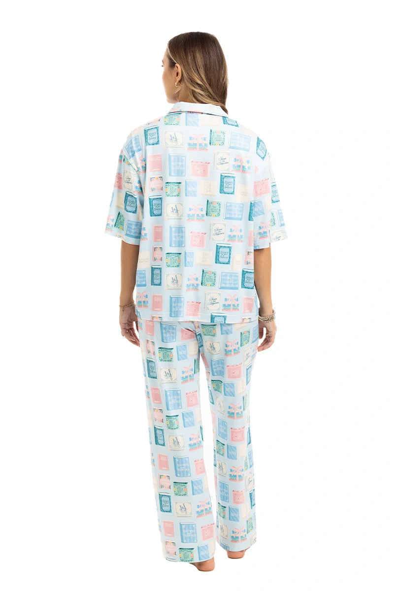 A woman stands barefoot facing away, wearing Katydids Book Club Matching Pajama Set in light blue and pink. The collared top and long pants feature rectangular block patterns—ideal for book lovers. White background.