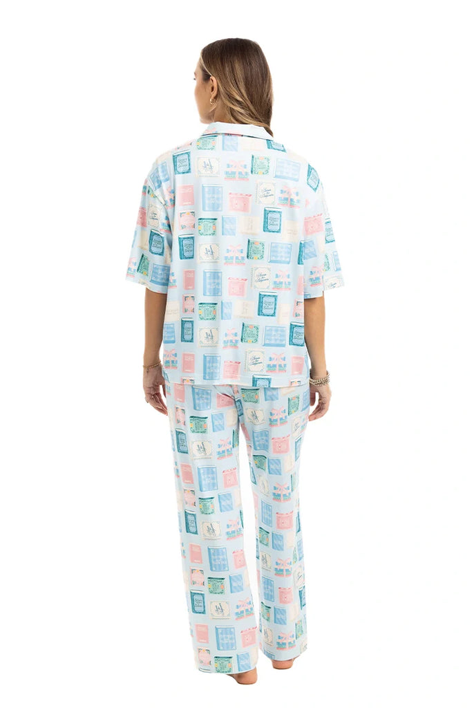 A woman stands barefoot facing away, wearing Katydids Book Club Matching Pajama Set in light blue and pink. The collared top and long pants feature rectangular block patterns—ideal for book lovers. White background.