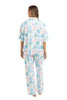 A woman stands barefoot facing away, wearing Katydids Book Club Matching Pajama Set in light blue and pink. The collared top and long pants feature rectangular block patterns—ideal for book lovers. White background.