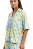 A woman with straight brown hair wears the Katydid Zesty Lemons Matching Pajama Pants Set—a light blue, short-sleeve loungewear outfit—smiling slightly and standing, perfect for relaxing comfortably at home.