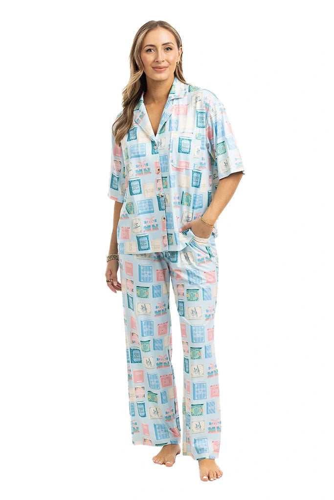 A woman models Katydids Book Club Matching Pajama Sets—light blue pants and a short-sleeve top with pastel book cover prints—standing barefoot against a white background.