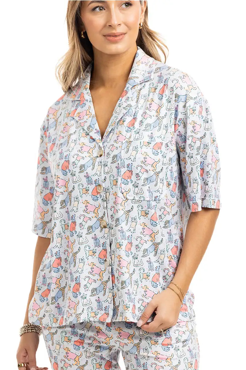 A woman with light brown hair, smiling softly, models the Katydid Posh Pets Preppy Pajama Pants—a colorful, playful animal print sleepwear perfect for pet lovers. She wears the matching short-sleeve button-up top with her hand at her side.