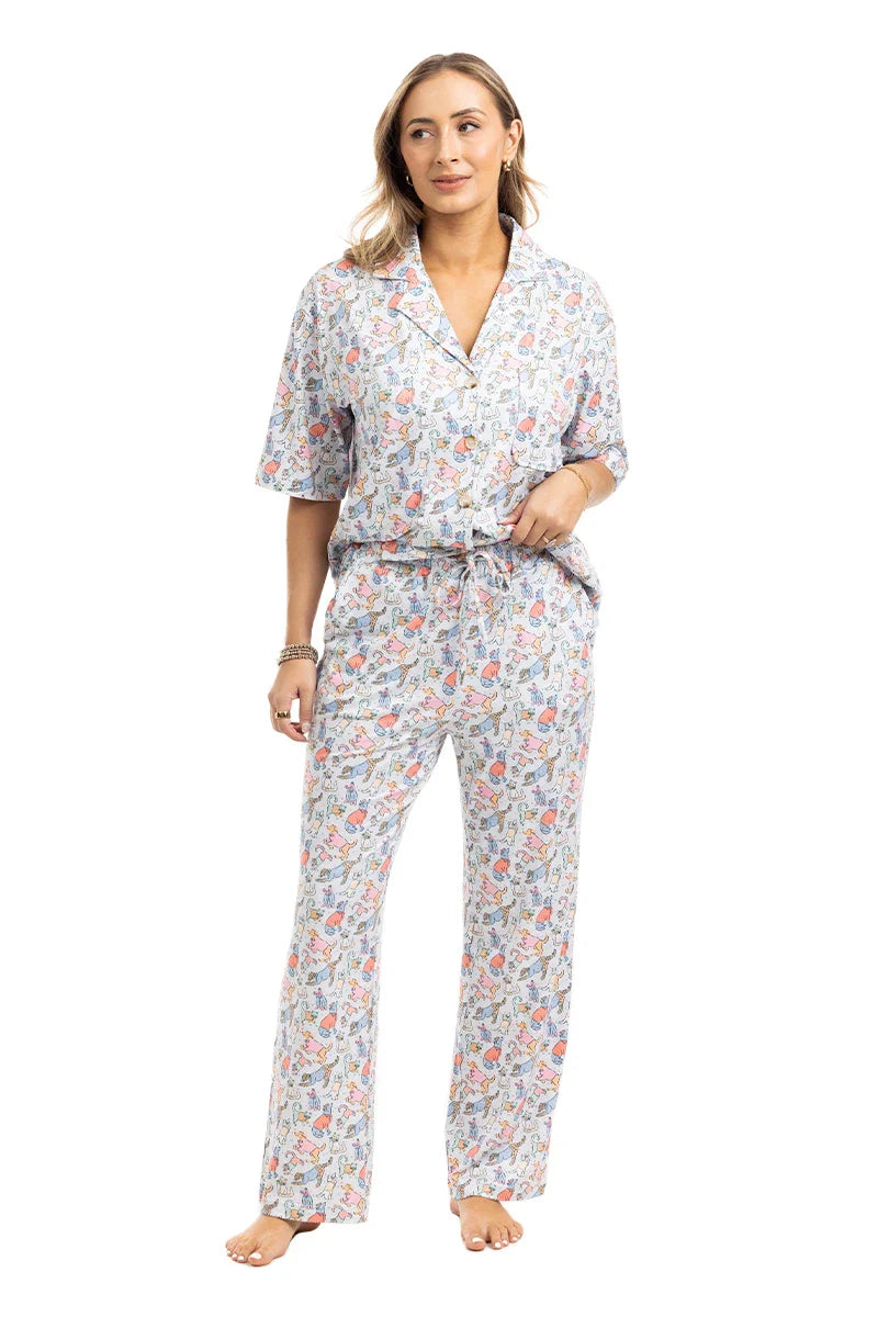A woman with light brown hair and a relaxed expression stands barefoot in Katydid’s Posh Pets Preppy Pajama Pants—a colorful floral, pet lover set featuring a short-sleeve button-up top and drawstring pants, perfect for animal enthusiasts.