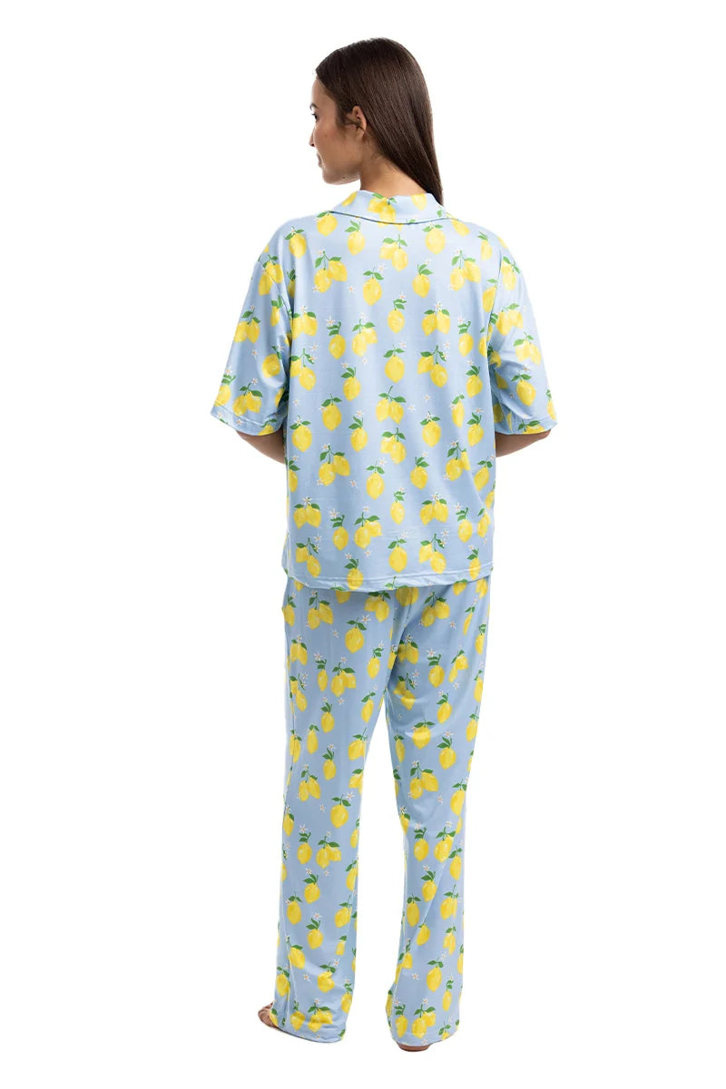 A woman, barefoot against a white background, wears the Katydid Zesty Lemons Matching Pajama Pants Set—a comfy womens light blue pajama with playful lemon print, featuring a short-sleeve top and long pants.