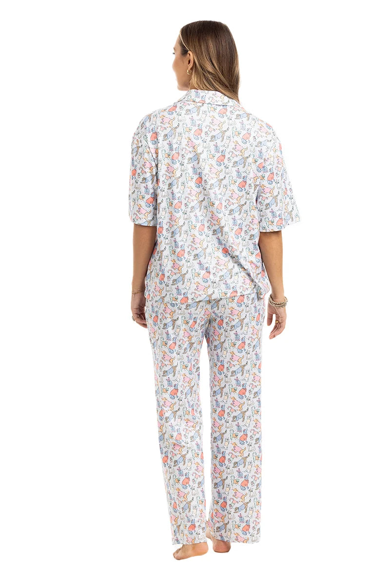 A woman stands barefoot, facing away, wearing Katydids Posh Pets Preppy Pajama Pants with a colorful floral pattern on white. This animal lover’s pajama set includes a short-sleeve top and full-length pants. The background is plain white.