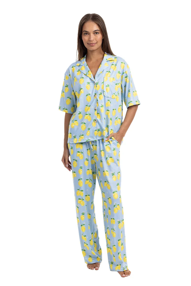 A woman wears the Katydid Zesty Lemons Matching Pajama Pants Set, featuring a light blue short-sleeve button-up shirt and long, loose pants with a bright yellow lemon print. She is barefoot and smiles slightly.