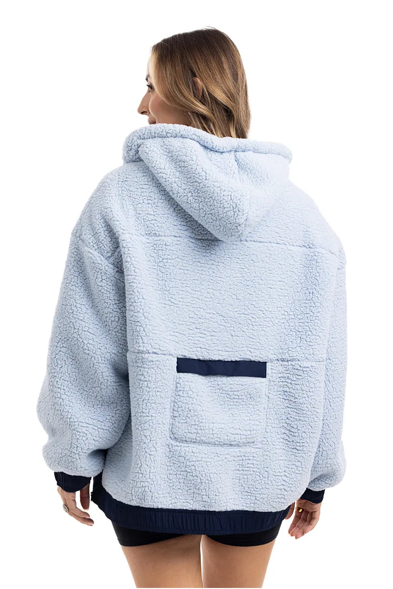 A person with shoulder-length hair is wearing the Katydid Light Blue Two-Toned Sherpa Fleece Pullover, featuring a cozy hood, dark blue trim, and a rectangular pocket on the lower back. They are facing away from the camera.