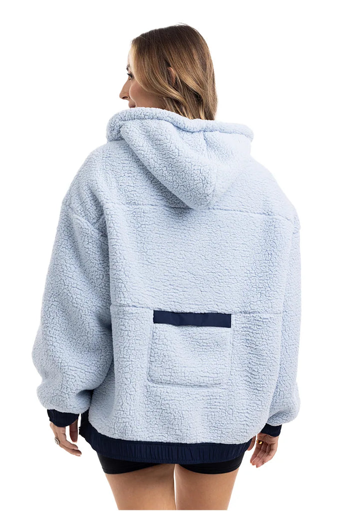 A person with shoulder-length hair is wearing the Katydid Light Blue Two-Toned Sherpa Fleece Pullover, featuring a cozy hood, dark blue trim, and a rectangular pocket on the lower back. They are facing away from the camera.
