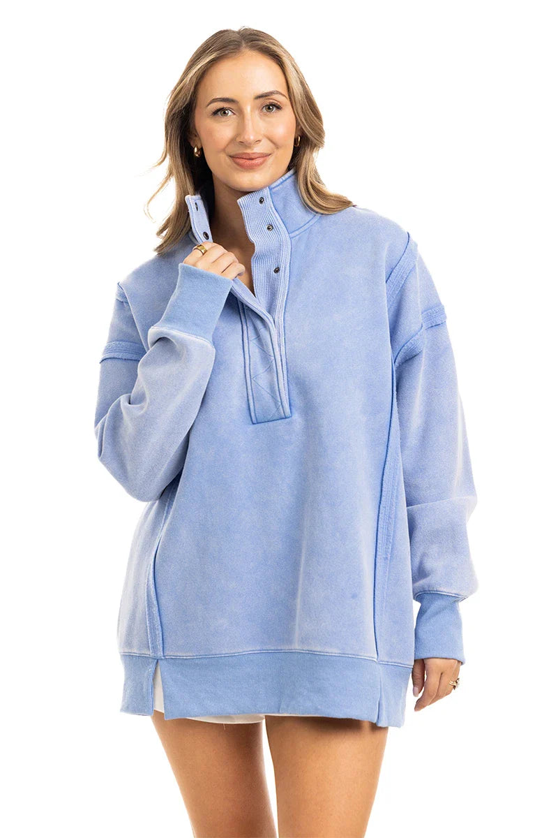 A woman with light brown hair smiles in the Katydid Light Blue Vintage Wash Snap-Neck Pullover, featuring a soft garment-washed finish and high collar, standing against a plain white background.