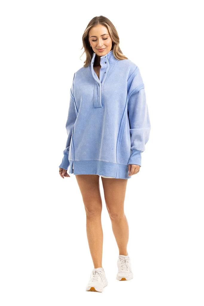 A woman in the Katydid Light Blue Vintage Wash Snap-Neck Pullover, styled with white sneakers, stands against a plain white background, smiling and looking down.