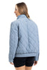 A woman with light brown hair models the Katydid Steel Blue Sporty Diamond Quilted Womens Jacket, paired with black shorts, facing away and turning slightly left against a white background.