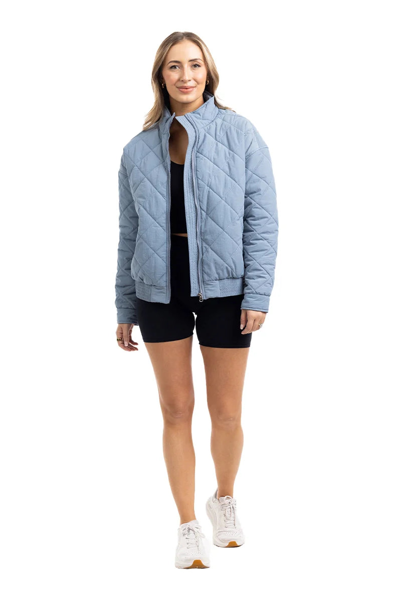 A woman in a Katydid Steel Blue Sporty Diamond Quilted Womens Jacket, black shorts, and white sneakers smiles against a white background.