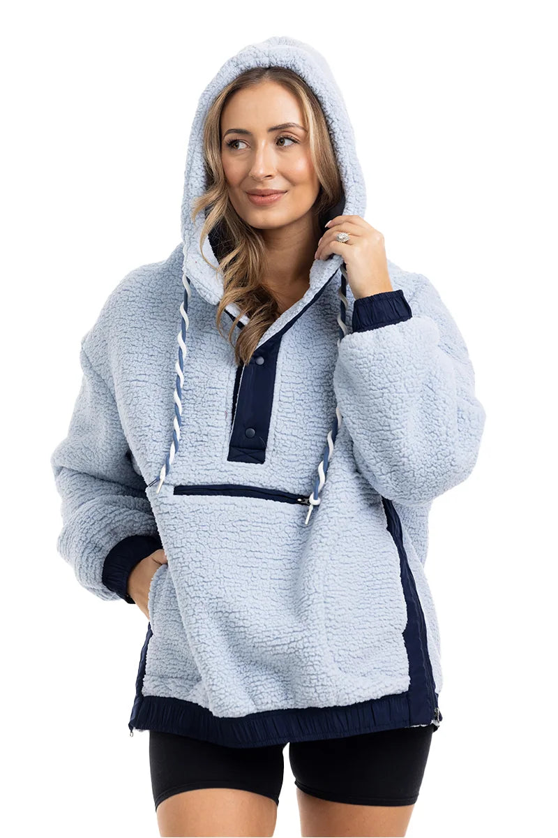 A woman wears the Katydid Light Blue Two-Toned Sherpa Fleece Pullover with navy accents and a front pocket, paired with black shorts. Smiling warmly, she touches the hood, showcasing the comfort and style of this cozy outerwear.