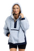 A woman wears the Katydid Light Blue Two-Toned Sherpa Fleece Pullover with navy accents and a front pocket, paired with black shorts. Smiling warmly, she touches the hood, showcasing the comfort and style of this cozy outerwear.