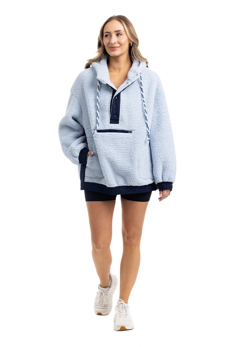 A woman in a Katydid Light Blue Two-Toned Sherpa Fleece Pullover with a front pocket, navy shorts, and white sneakers walks forward, smiling against a plain white background.