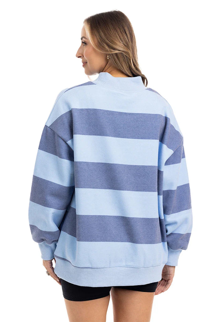 A woman stands facing away, wearing the Katydid Light Blue and Navy Striped Mockneck Sweatshirt with black shorts—ideal for fall layering. Her hair is loose as she smiles slightly, glancing to the side.