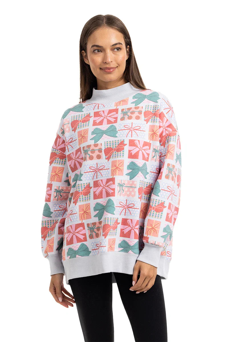 A woman with straight brown hair models the Katydid Wrapped Presents with Bows Mockneck Pullover, featuring colorful gift box patterns, paired with black leggings and posed against a white background.
