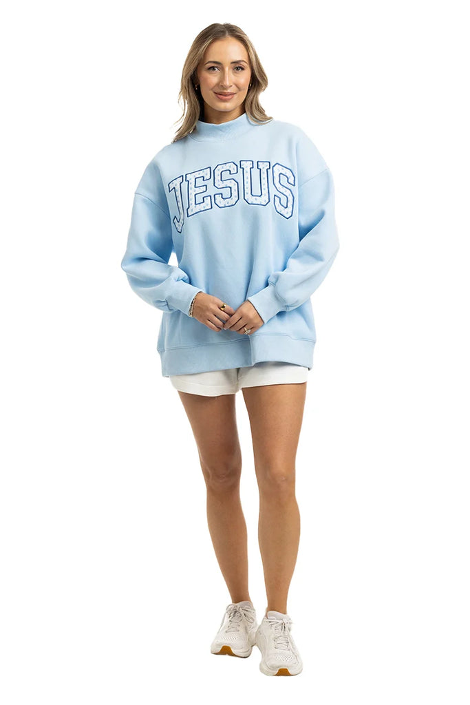 A woman smiles in the Katydid JESUS Mockneck sweatshirt, paired with white shorts and sneakers—a stylish pick for Christian apparel—set against a simple white background.