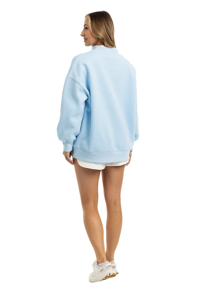 A woman faces away, wearing the Katydid JESUS Mockneck sweatshirt in light blue, paired with white shorts and sneakers. Her long light brown hair is visible against a plain white background.