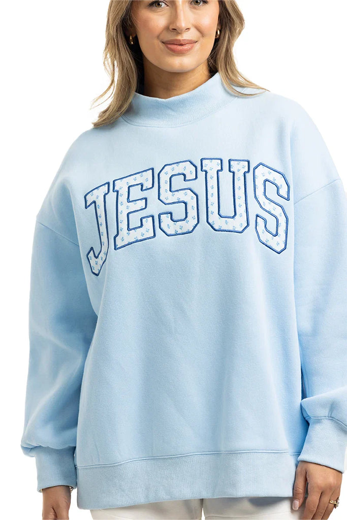 A woman models the Katydid JESUS Mockneck, a light blue embroidered sweatshirt featuring bold white and blue JESUS lettering across the chest—ideal stylish Christian apparel for any wardrobe.