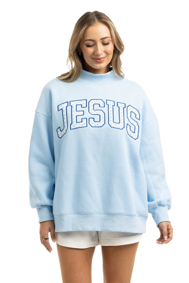 A woman wears the Katydid JESUS Mockneck in light blue with bold white letters, paired with white shorts. She stands against a plain white background, looking down and smiling—ideal Christian apparel for a relaxed, stylish look.
