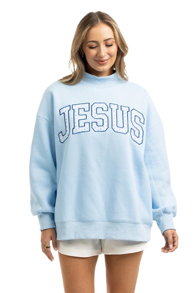 A woman wears the Katydid JESUS Mockneck in light blue with bold white letters, paired with white shorts. She stands against a plain white background, looking down and smiling—ideal Christian apparel for a relaxed, stylish look.