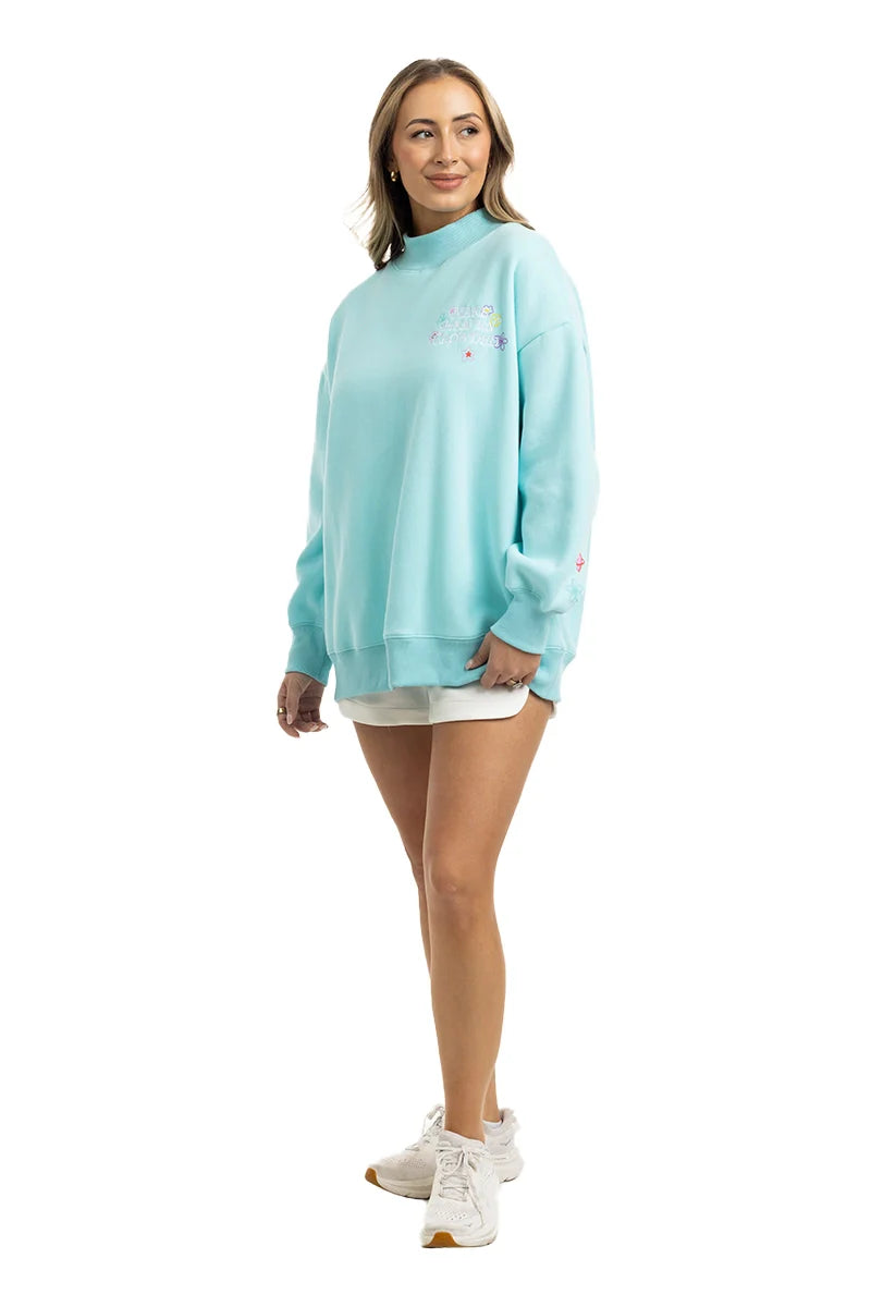A woman smiles, wearing a light blue Katydid Make Heaven Crowded Womens Mockneck Top with white shorts and sneakers, set against a plain white background.