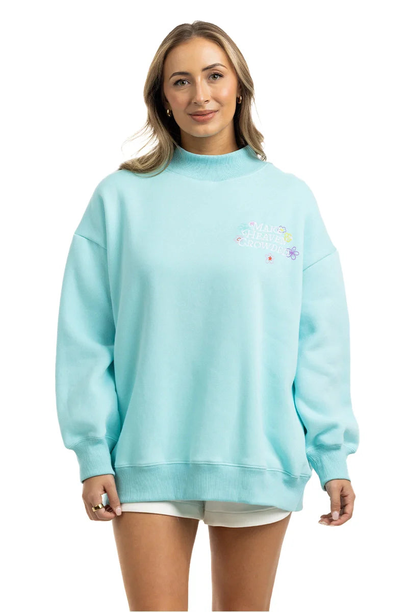 A woman in a light blue Katydid Make Heaven Crowded Women’s Mockneck Top with small colorful text and floral graphics on the chest, paired with white shorts, stands smiling against a white background.