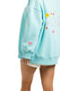 A person in a light blue, oversized Katydid Make Heaven Crowded Womens Mockneck Top with colorful embroidered back graphics, paired with white shorts. The image is cropped to show only the lower face and body from the side.