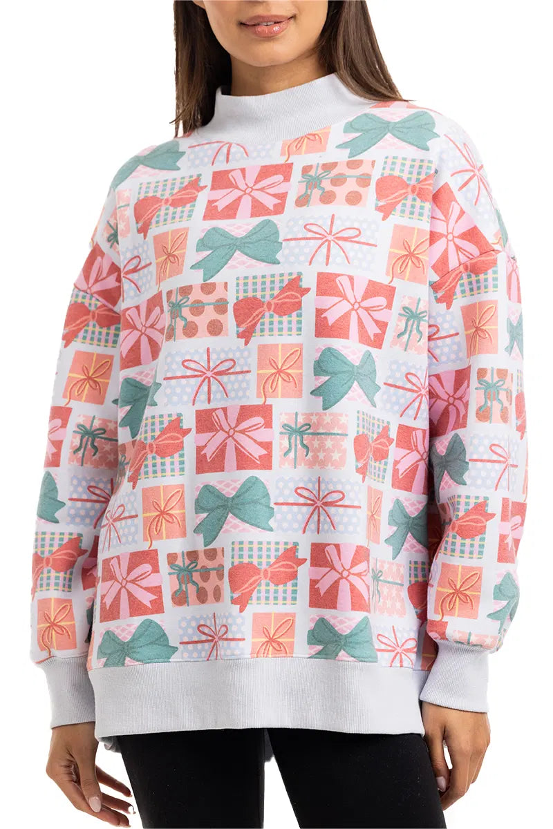 A woman stands with her hands at her sides, wearing the Katydid Wrapped Presents with Bows mockneck pullover—a holiday sweatshirt featuring a colorful pattern of gift boxes and bows in red, pink, green, and white.