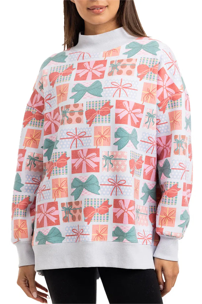 A woman stands with her hands at her sides, wearing the Katydid Wrapped Presents with Bows mockneck pullover—a holiday sweatshirt featuring a colorful pattern of gift boxes and bows in red, pink, green, and white.