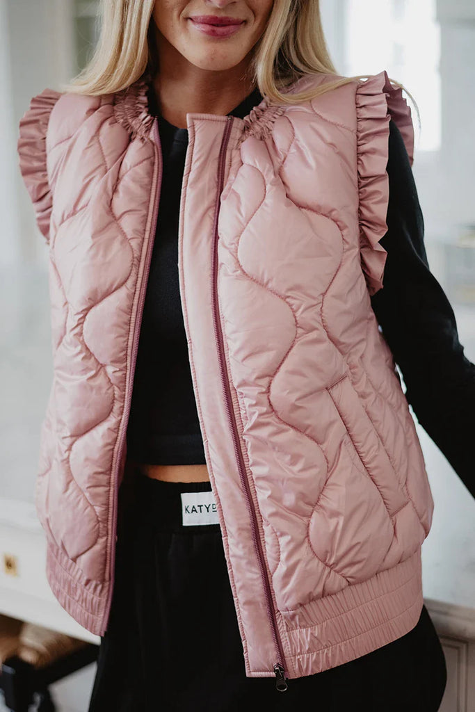 A woman indoors smiles slightly, wearing the Katydid Blush Metallic Puffy Vest over a black top. The lightweight vest features quilted fabric, a feminine ruffle detail, subtle sheen, and front zipper. Only her torso and lower face are visible.