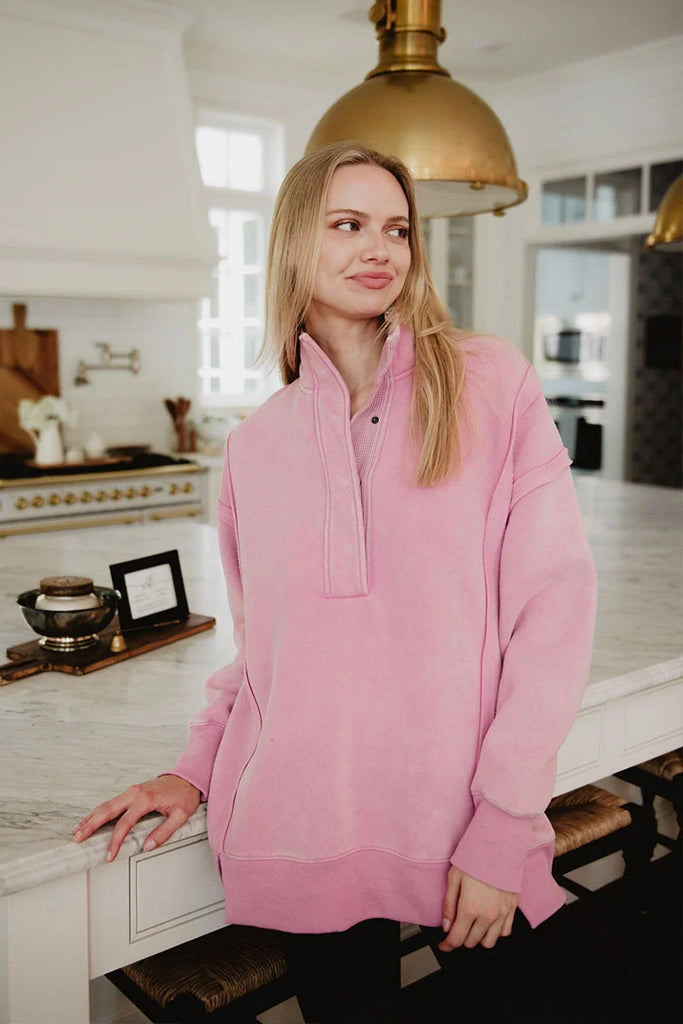 A woman in a Katydid Pink Vintage Wash Pullover for Ladies stands in a bright, modern kitchen, smiling softly as she looks to the side with one hand on a marble countertop.