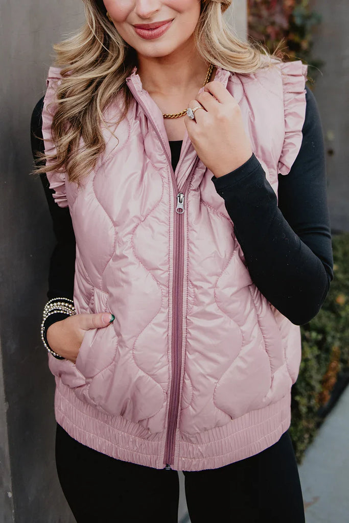 A woman smiles in a Katydid Blush Metallic Puffy Vest with ruffled shoulders, layered over a black long-sleeve top. One hand is in her pocket while the other touches her necklace.