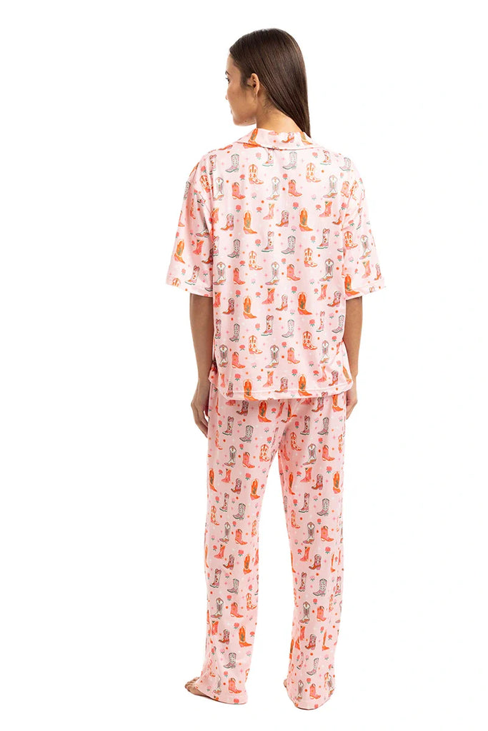 A woman faces away, wearing the Katydid Womens Boots  Roses PJ Set— a pink short-sleeve, loose pants pajama set for women featuring a colorful Western print, styled as cozy cowgirl loungewear against a plain white background.