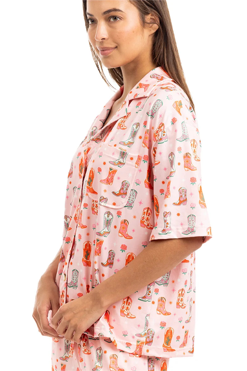 A woman stands sideways, smiling gently in the Katydid Womens Boots  Roses PJ Set—a cute, loose-fitting pajama with a colorful cowboy boot pattern and short sleeves for comfy cowgirl style.
