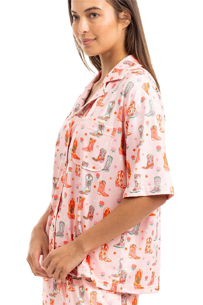 A woman stands sideways, smiling gently in the Katydid Womens Boots  Roses PJ Set—a cute, loose-fitting pajama with a colorful cowboy boot pattern and short sleeves for comfy cowgirl style.