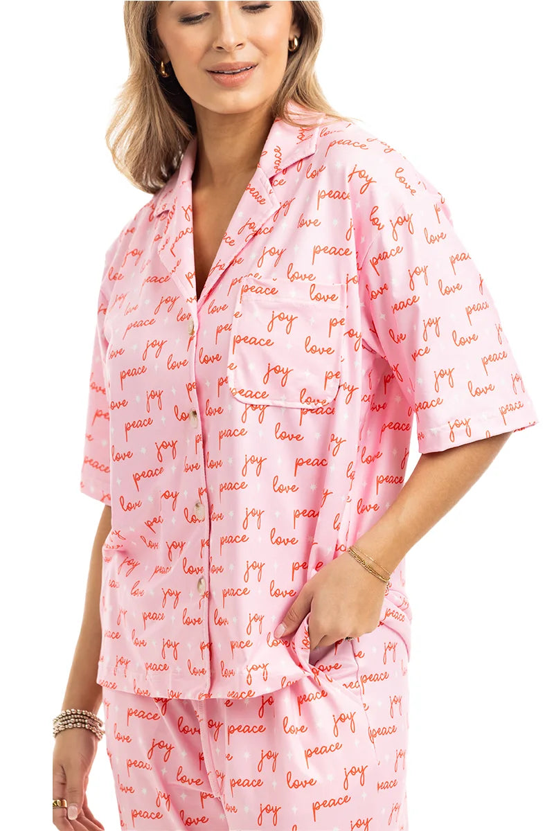 A woman beams in the Katydid Love Joy  Peace Preppy PJ Set—pink with red “love, joy, peace” print. The holiday pajamas include a short-sleeve collared button top with chest pocket and comfy pants with side pockets.